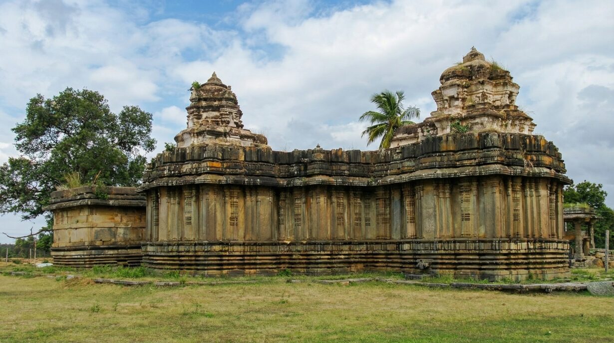Ancient stone temple architecture surrounded by greenery under a cloudy sky, featuring intricately carved pillars