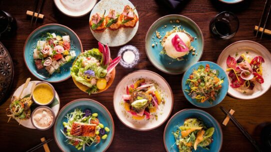 Overhead shot of vibrant Asian-fusion small plates revealing the real story behind the food through seared fish, bao, and floral salads on a dark table.