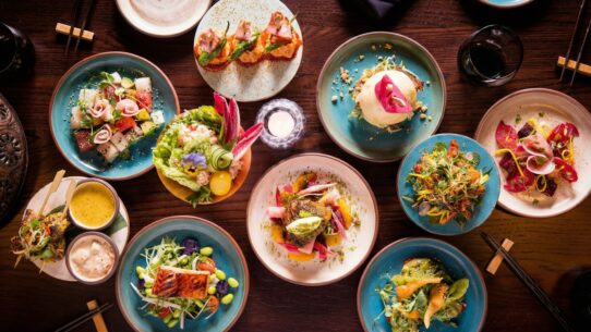 Overhead shot of vibrant Asian-fusion small plates revealing the real story behind the food through seared fish, bao, and floral salads on a dark table.
